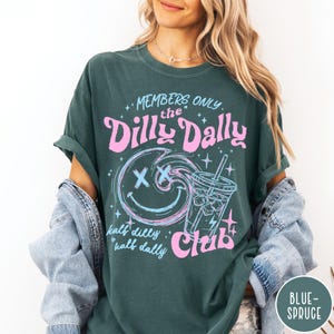 May include: Dark teal t-shirt with a graphic design featuring the text "MEMBERS ONLY the Dilly Dally Club." The design includes a smiling face, a beverage, and the words "half dilly half dally." The shirt is a relaxed fit.
