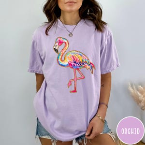 May include: A light purple t-shirt featuring a colorful flamingo design. The flamingo has a rainbow-colored body with gold glitter accents and is standing on one leg. The word "ORCHID" is in a purple circle in the bottom right corner.