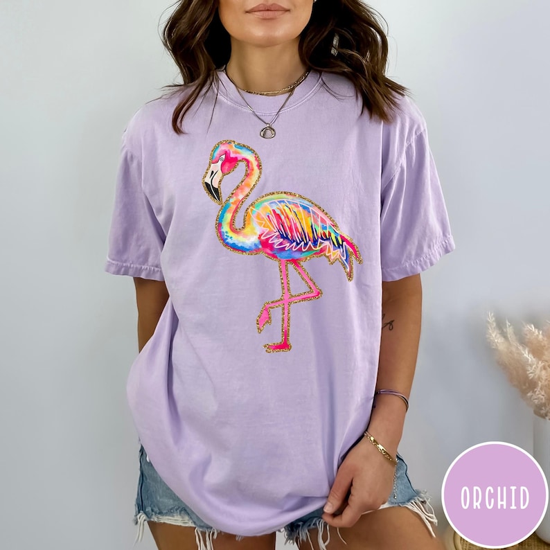 May include: A light purple t-shirt featuring a colorful flamingo design. The flamingo has a rainbow-colored body with gold glitter accents and is standing on one leg. The word "ORCHID" is in a purple circle in the bottom right corner.