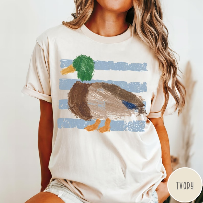 May include: An ivory short-sleeve t-shirt featuring a graphic print of a mallard duck with a green head, brown body, and orange feet. The duck is layered over a background of horizontal, distressed light blue stripes across the center chest.
