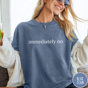 May include: A blue-grey crew neck sweatshirt with the words "immediately no" in white. The sweatshirt has a relaxed fit and is part of the "Blue Jean" collection. The model is wearing sunglasses and a necklace.