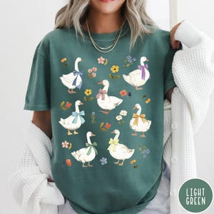 K&ouml;nnte beinhalten: Ein hellgr&uuml;nes T-Shirt mit einem Muster aus wei&szlig;en G&auml;nsen mit bunten Schleifen und floralen Akzenten.
