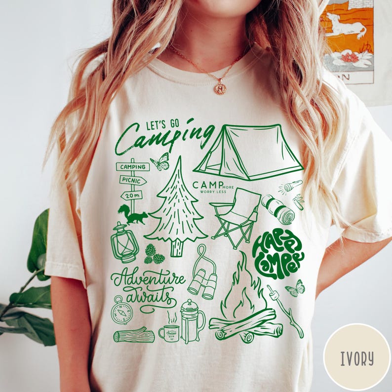 Może przedstawiać: Kremowa koszulka z zieloną grafiką o tematyce kempingowej. Projekt zawiera tekst "Let's Go Camping" i "Adventure Awaits", a także ilustracje namiotu, ogniska i innych niezbędnych element&oacute;w kempingowych.