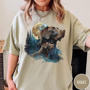 May include: Khaki-colored t-shirt featuring a watercolor illustration of two Labrador Retrievers under a full moon. The design includes a forest scene with blue and green accents. The shirt has short sleeves and a relaxed fit.
