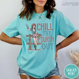 May include: A turquoise t-shirt with the text "CHILL THE FOURTH OUT" and a graphic of a woman in a patriotic outfit holding a firework. The design is in red, white, and blue. The shirt is a relaxed fit.