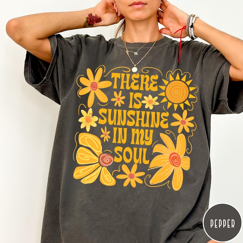 May include: Dark gray t-shirt with a yellow floral design and the words 'THERE IS SUNSHINE IN MY SOUL' in a retro font. The design includes stylized yellow flowers and a sun graphic. The shirt has a vintage, slightly faded look.
