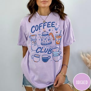 May include: A light purple t-shirt with a coffee-themed graphic. The design includes a coffee pot, cups, and the words "Coffee Club" in blue. The shirt has a relaxed fit and short sleeves.