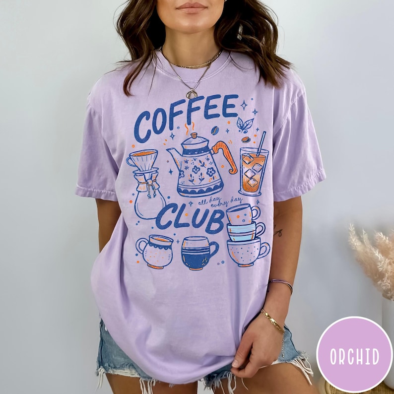 May include: A light purple t-shirt with a coffee-themed graphic. The design includes a coffee pot, cups, and the words "Coffee Club" in blue. The shirt has a relaxed fit and short sleeves.