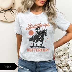 May include: A grey t-shirt with a graphic of a woman riding a horse. The text "Saddle up, Buttercup!" is printed on the shirt.