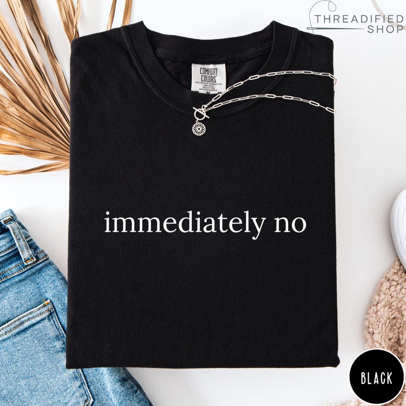 May include: Black t-shirt with the words "immediately no" in white. A silver necklace with a sun-shaped pendant rests on the shirt. The shirt has a Comfort Colors tag and a "Threadified Shop" logo.