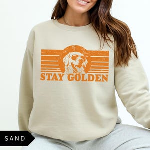 Puede incluir: Una sudadera beige con un gr&aacute;fico naranja de un golden retriever con gafas de sol y el texto "Stay Golden".