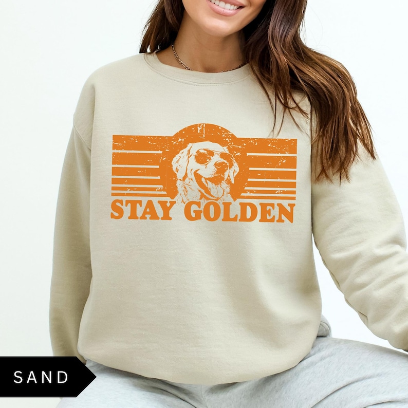 Puede incluir: Una sudadera beige con un gr&aacute;fico naranja de un golden retriever con gafas de sol y el texto "Stay Golden".