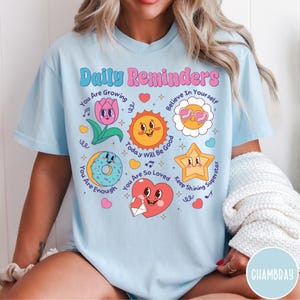 May include: Light blue t-shirt with the words "Daily Reminders" and various cartoon graphics, including a flower, sun, donut, star, and heart. The shirt has positive affirmations like "You Are Growing" and "You Are Enough".