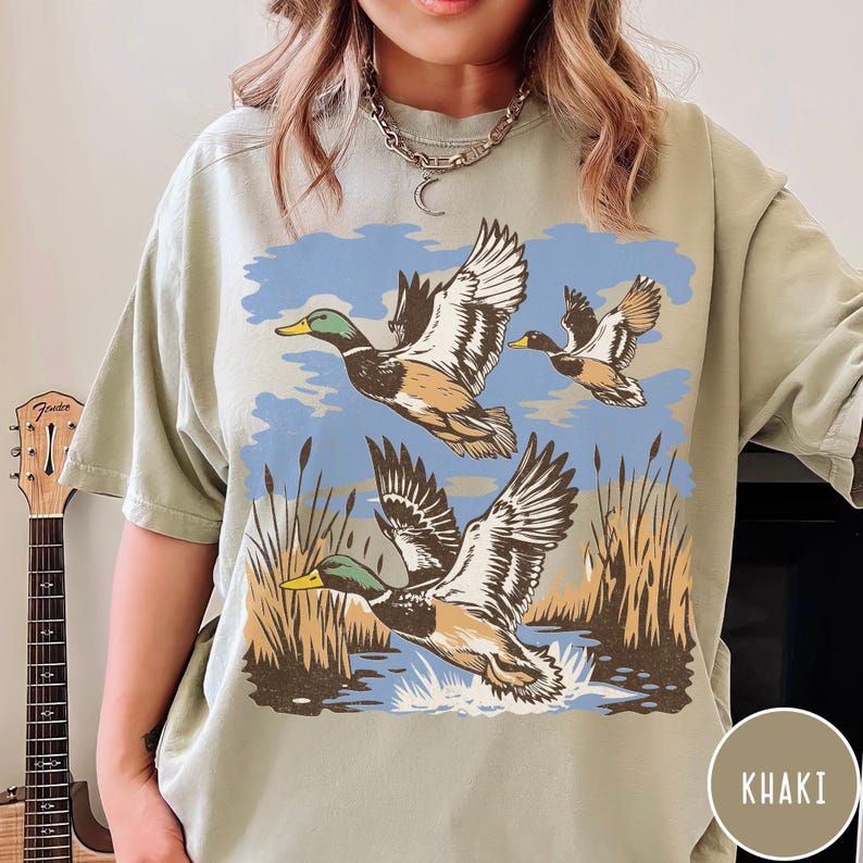 May include: Khaki-coloured t-shirt with a graphic of three ducks flying over a marsh. The design includes blue sky, water, and reeds. The shirt has a relaxed fit.