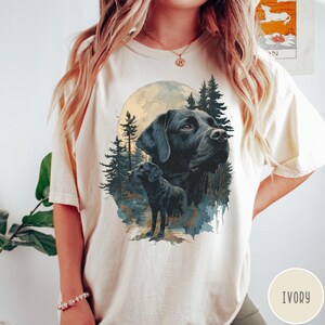 May include: Ivory-colored t-shirt featuring a detailed illustration of a black Labrador Retriever. The design includes a full moon, pine trees, and a smaller image of the dog. The shirt has a relaxed fit and short sleeves.