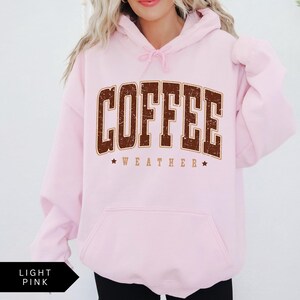 Puede incluir: Una sudadera con capucha rosa claro con un gr&aacute;fico marr&oacute;n que dice "COFFEE WEATHER" con estrellas a cada lado del texto.