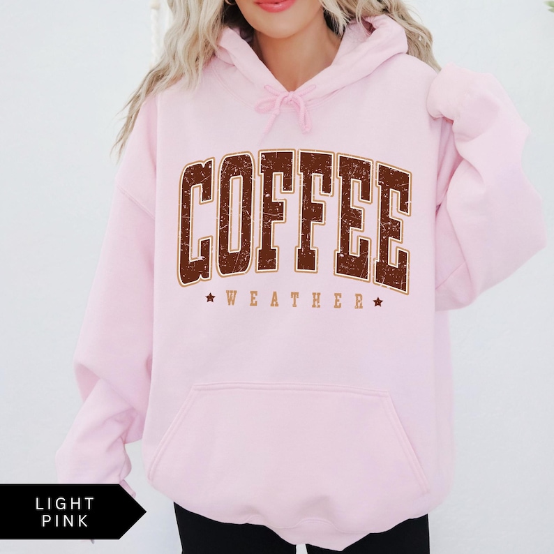 Puede incluir: Una sudadera con capucha rosa claro con un gr&aacute;fico marr&oacute;n que dice "COFFEE WEATHER" con estrellas a cada lado del texto.