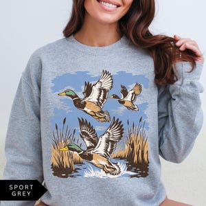 May include: Sport grey sweatshirt featuring a graphic of three ducks in flight over water and reeds. The ducks have brown, white, and green details against a blue sky. The sweatshirt has the words "SPORT GREY" printed on the bottom left.