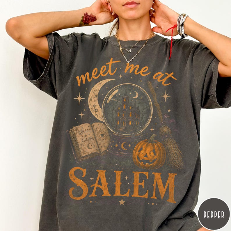 May include: A charcoal gray t-shirt featuring a graphic print with the text "meet me at SALEM". The design includes a crystal ball containing a house, an open book titled "SPELLS & CHARMS", a crescent moon, a broomstick, and an orange pumpkin.