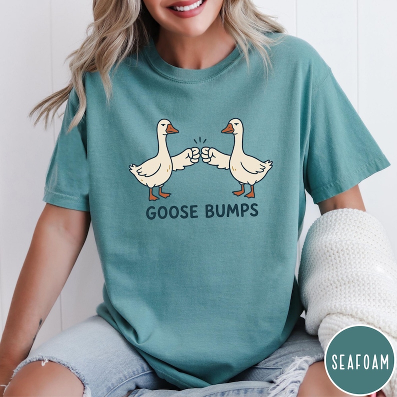 Puede incluir: Una camiseta de manga corta color verde espuma de mar con un gr&aacute;fico de dos gansos blancos chocando los pu&ntilde;os. Debajo de los gansos, el texto "GOOSE BUMPS" aparece en letras may&uacute;sculas de color verde oscuro. Tiene cuello redondo.