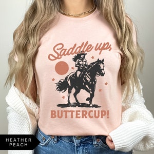 May include: A pink t-shirt with a vintage-style graphic of a woman riding a horse. The text on the shirt reads "Saddle up, Buttercup!"