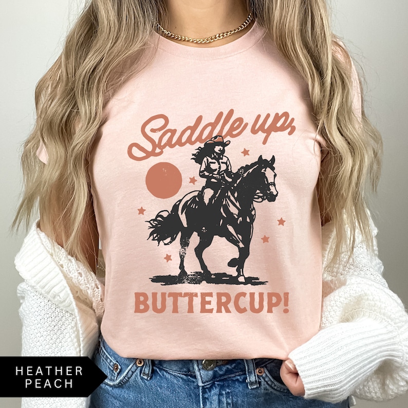 May include: A pink t-shirt with a vintage-style graphic of a woman riding a horse. The text on the shirt reads "Saddle up, Buttercup!"
