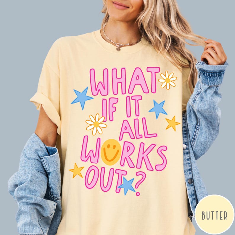 May include: A butter yellow short-sleeve t-shirt featuring the pink text WHAT IF IT ALL WORKS OUT? with a yellow smiley face replacing the letter O. The design includes light blue stars, yellow stars, and white daisies with yellow centers.