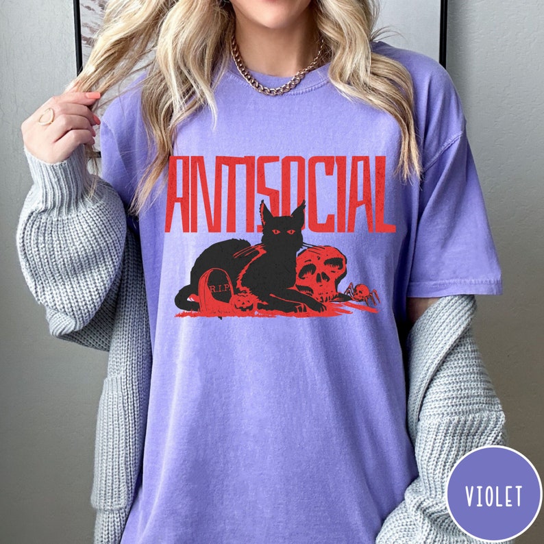 Może przedstawiać: Fioletowy T-shirt z napisem "ANTISOCIAL" w czerwonych literach. Na koszulce znajduje się czarny kot z czerwonymi oczami siedzący obok czaszki i nagrobka z napisem "RIP".