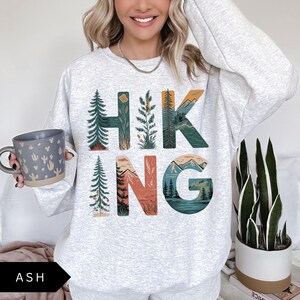 Puede incluir: Sudadera gris ceniza con la palabra "HIKING" en un dise&ntilde;o tem&aacute;tico de la naturaleza. Las letras est&aacute;n rellenas de im&aacute;genes de &aacute;rboles, monta&ntilde;as y agua. Una taza gris con un dise&ntilde;o de cactus se sostiene en la mano izquierda.