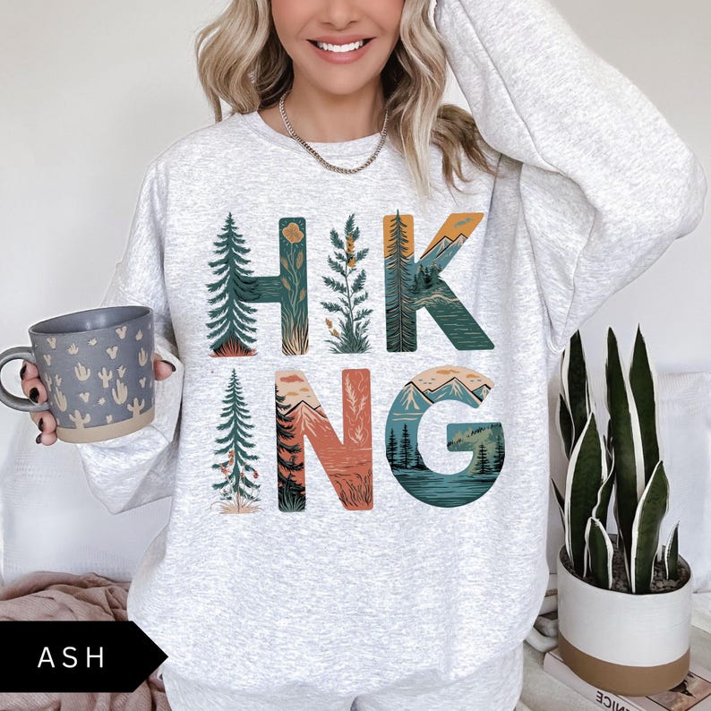 Puede incluir: Sudadera gris ceniza con la palabra "HIKING" en un dise&ntilde;o tem&aacute;tico de la naturaleza. Las letras est&aacute;n rellenas de im&aacute;genes de &aacute;rboles, monta&ntilde;as y agua. Una taza gris con un dise&ntilde;o de cactus se sostiene en la mano izquierda.