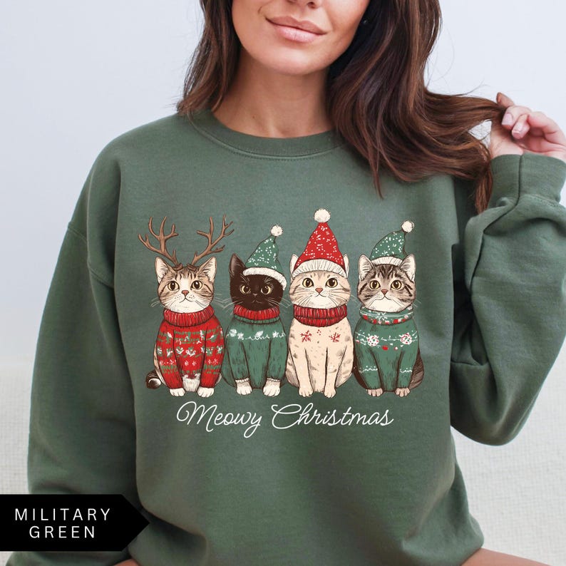 May include: Military green sweatshirt featuring a graphic of four cats dressed in Christmas sweaters and hats. The cats are wearing red and green sweaters and hats, with the text "Meowy Christmas" below the cats.