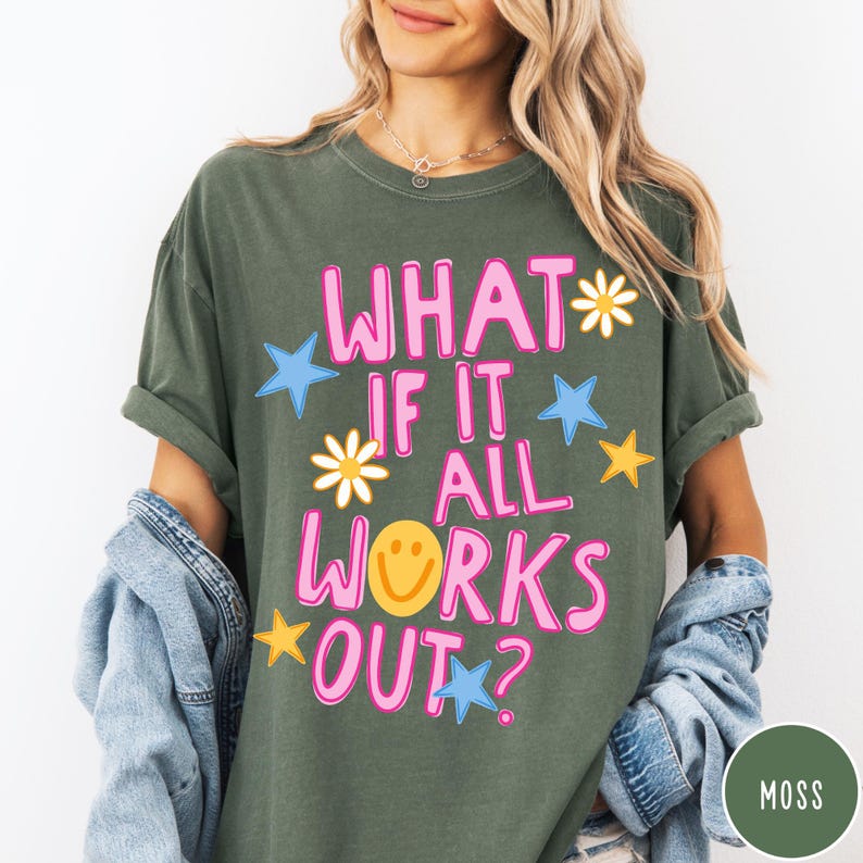 May include: A moss green short-sleeve t-shirt featuring the pink text "WHAT IF IT ALL WORKS OUT?" with a yellow smiley face replacing the letter O. The design includes scattered blue stars, yellow stars, and white daisy flower graphics.