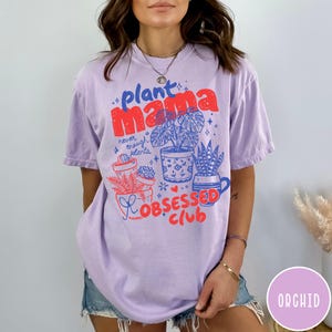 Puede incluir: Camiseta lila claro con las palabras "plant mama" en rojo y azul, junto con ilustraciones de plantas en macetas. Tambi&eacute;n aparece el texto "Obsessed Club". La camiseta es de corte holgado.