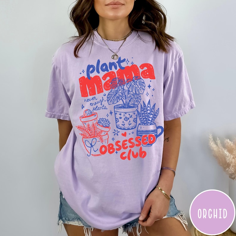 Puede incluir: Camiseta lila claro con las palabras "plant mama" en rojo y azul, junto con ilustraciones de plantas en macetas. Tambi&eacute;n aparece el texto "Obsessed Club". La camiseta es de corte holgado.