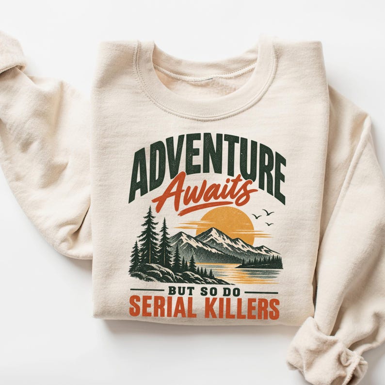 May include: A cream-colored crewneck sweatshirt featuring a graphic print of a mountain landscape with pine trees, a sun, and birds. The text reads ADVENTURE Awaits BUT SO DO SERIAL KILLERS in dark green and orange lettering across the chest.
