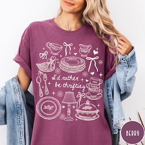 May include: Berry-coloured t-shirt featuring a white graphic of vintage kitchenware, such as plates and teacups, and a duck. The text "I'd rather be thrifting" is printed on the shirt. The sleeves are rolled up.