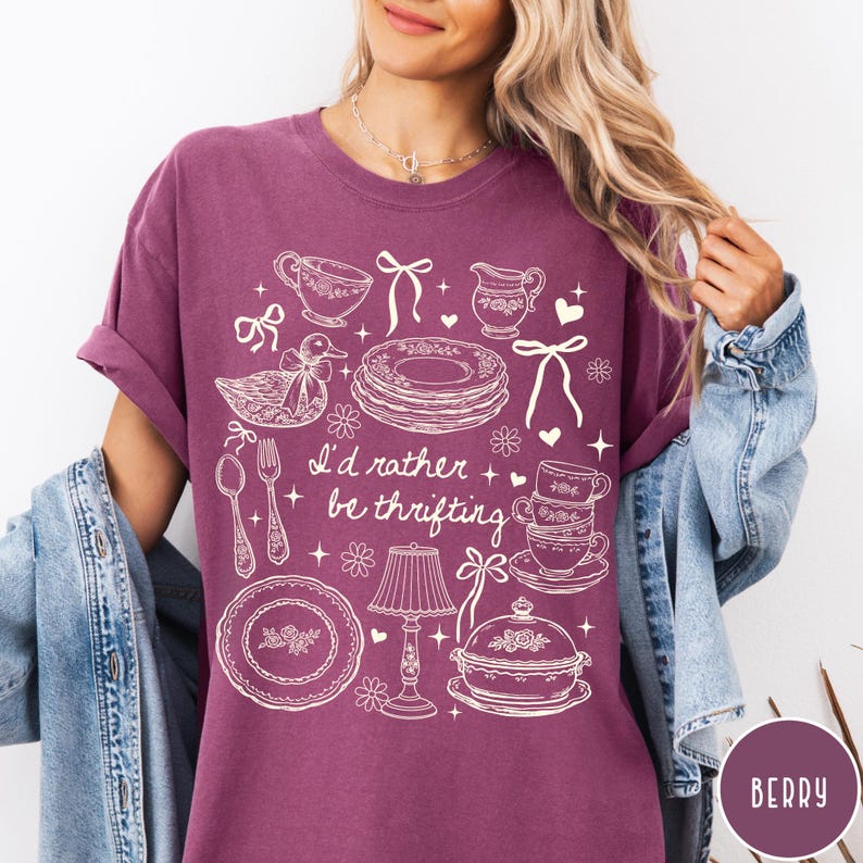 May include: Berry-coloured t-shirt featuring a white graphic of vintage kitchenware, such as plates and teacups, and a duck. The text "I'd rather be thrifting" is printed on the shirt. The sleeves are rolled up.