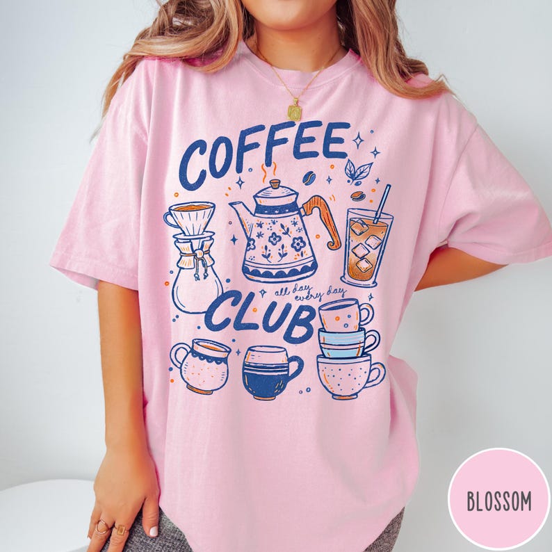 May include: A light pink t-shirt with a "Coffee Club" graphic in navy blue. The design includes coffee-related illustrations such as a teapot, coffee cups, and a glass of iced coffee. The shirt has short sleeves.