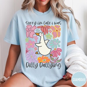 May include: A light blue t-shirt with a white goose and colorful flowers. The text "Sorry I'm late I was dilly dallying" is printed on the shirt.