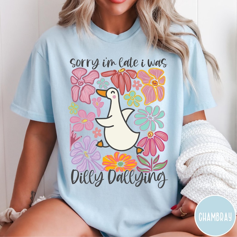 May include: A light blue t-shirt with a white goose and colorful flowers. The text "Sorry I'm late I was dilly dallying" is printed on the shirt.