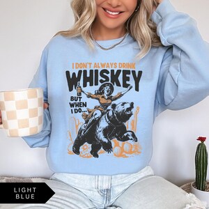 May include: Light blue sweatshirt with the text "I don't always drink whiskey, but when I do..." and a graphic of a woman riding a bear while holding a bottle of whiskey.