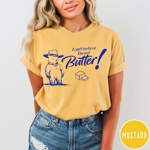 May include: A yellow t-shirt with a cartoon bear wearing a cowboy hat and the text "I can't believe I'm not Butter!" in blue. The shirt is on a model wearing blue jeans.