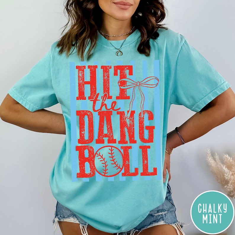 Pu&ograve; includere: T-shirt turchese chiaro con la scritta rossa "HIT the DANG BALL" e una grafica di baseball. Il testo &egrave; su uno sfondo a righe blu. La maglietta ha maniche corte e una vestibilit&agrave; ampia.
