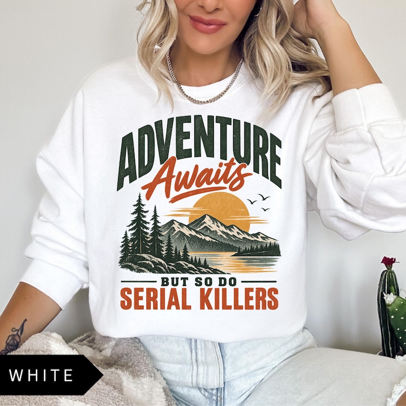 May include: A white long-sleeve sweatshirt featuring a graphic print of a mountain landscape with a sun, pine trees, and birds. The text reads ADVENTURE Awaits BUT SO DO SERIAL KILLERS in dark green and orange lettering across the front chest area.