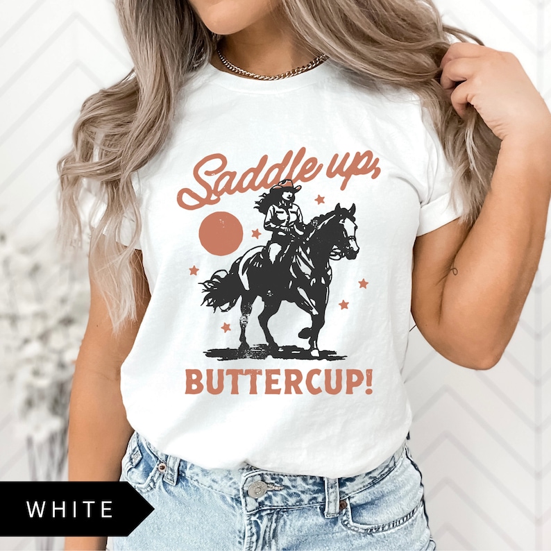May include: White t-shirt with a graphic of a woman riding a horse. The graphic includes the text "Saddle up, Buttercup!" in a brown font.