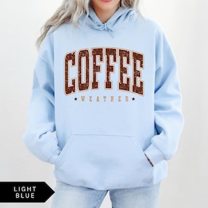 Puede incluir: Sudadera con capucha azul claro con un gr&aacute;fico marr&oacute;n desgastado que dice "Coffee Weather" con dos estrellas a cada lado del texto.