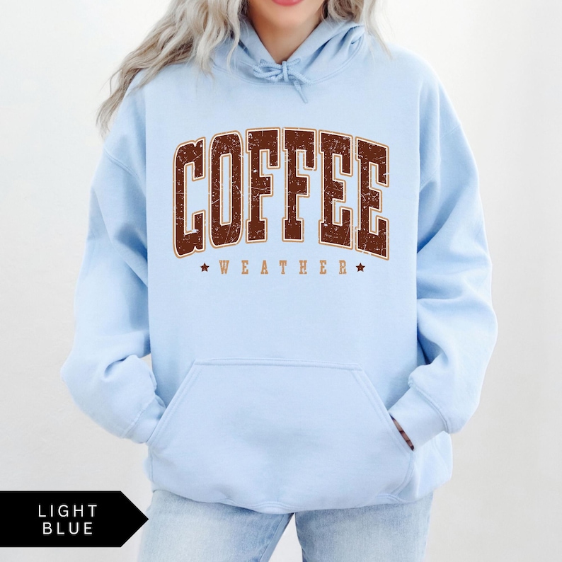 Puede incluir: Sudadera con capucha azul claro con un gr&aacute;fico marr&oacute;n desgastado que dice "Coffee Weather" con dos estrellas a cada lado del texto.