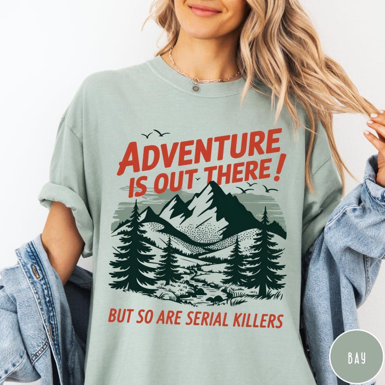 以下が含まれることがあります： 「ADVENTURE IS OUT THERE！」の赤い文字がプリントされた、山と森のグラフィックが特徴のライトグリーンのTシャツ。グラフィックの下には赤い文字で「BUT SO ARE SERIAL KILLERS」と書かれています。半袖のシャツです。
