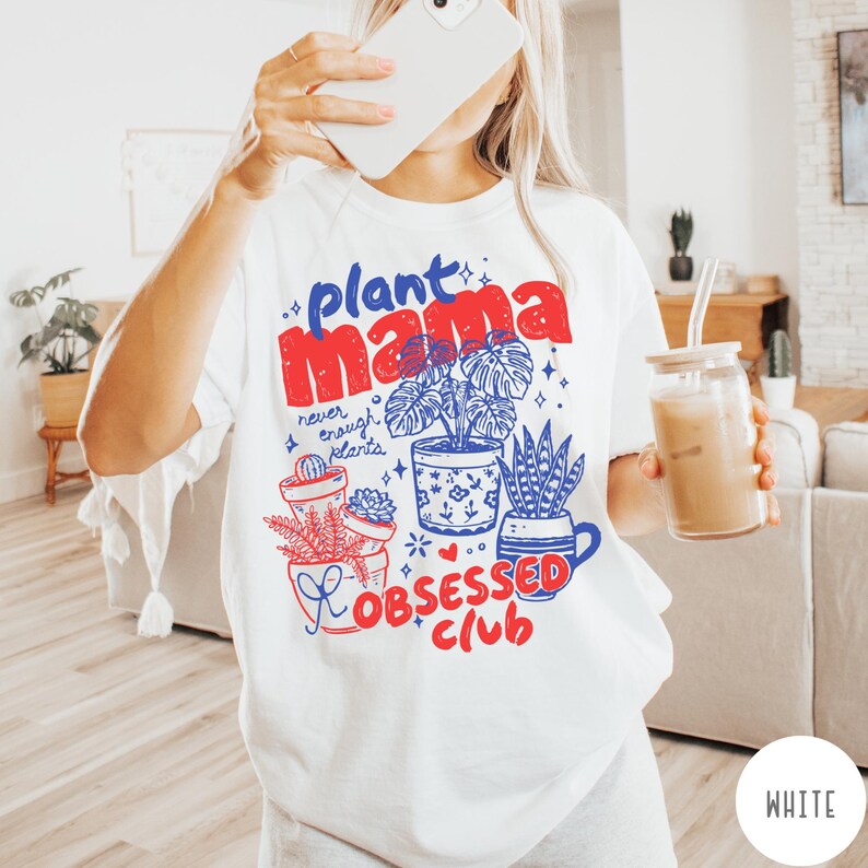 Puede incluir: Camiseta blanca con un dise&ntilde;o gr&aacute;fico colorido. El dise&ntilde;o presenta el texto "plant mama" en rojo y azul, junto con ilustraciones de plantas en macetas y las palabras "never enough plants" y "obsessed club".