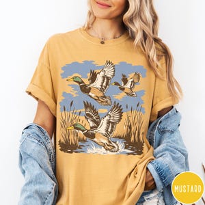 May include: Mustard-coloured t-shirt with a graphic of three ducks flying over water and reeds. The ducks are shown with brown, green, and white feathers against a blue sky. The shirt has a relaxed fit and rolled-up sleeves.